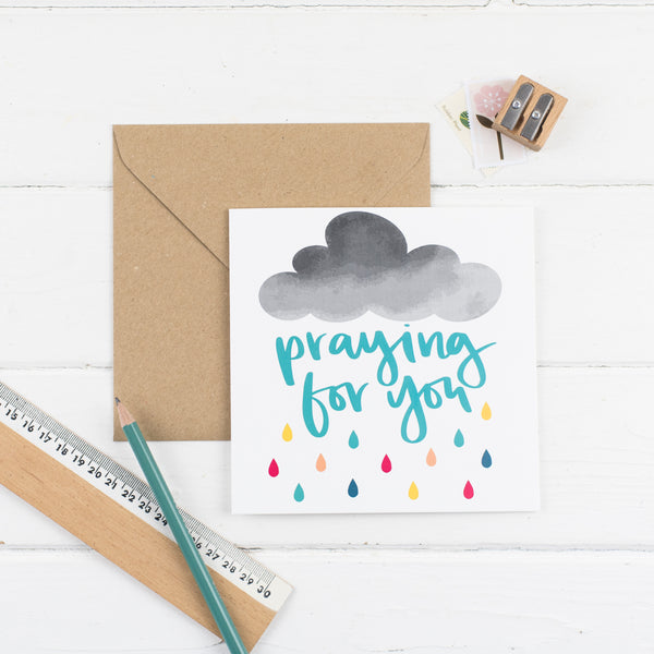 Praying for you square greeting card hand-lettered in teal amongst a cloud that is raining rainbow rain with kraft envelope